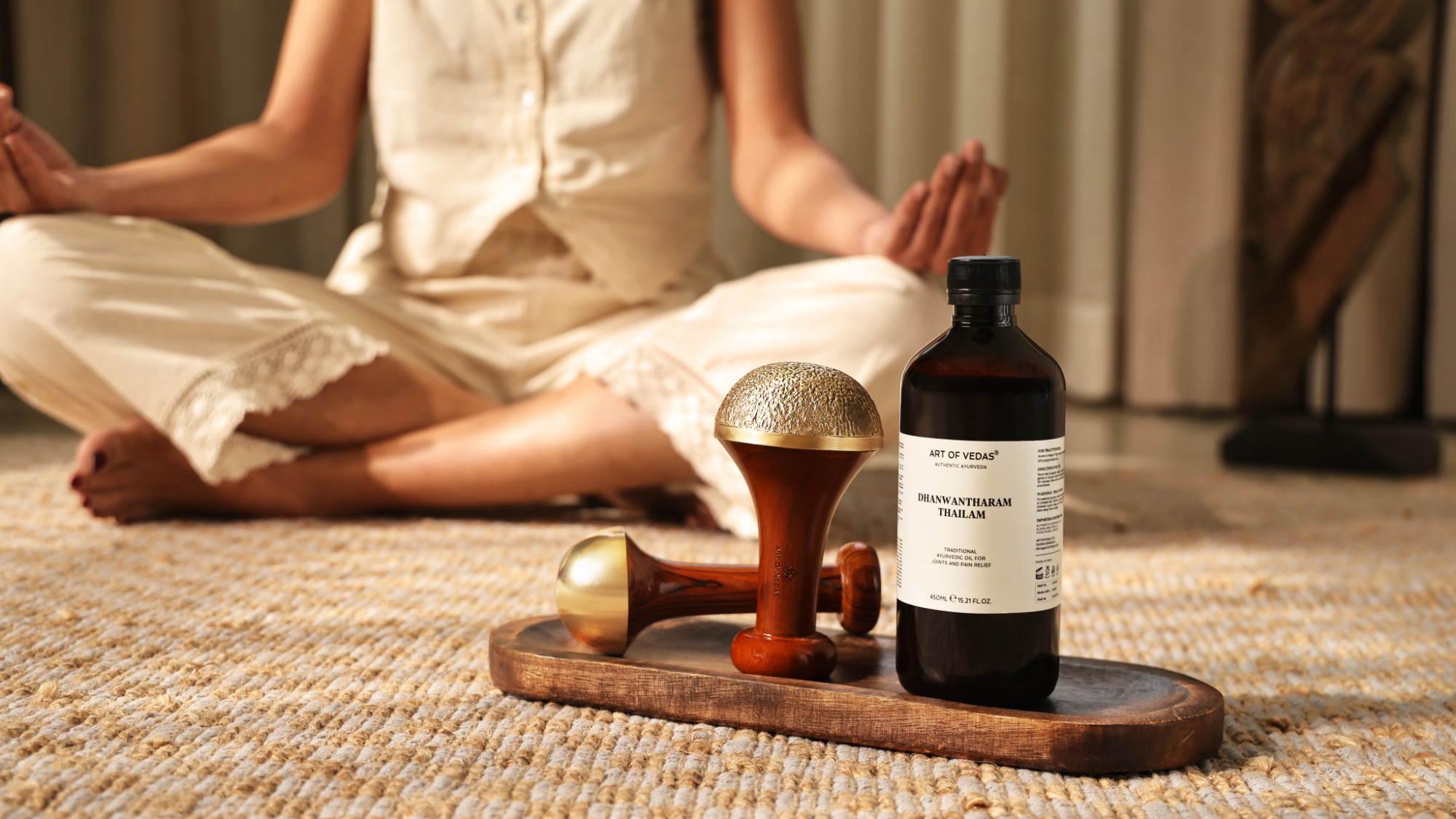 Zwei Kansa Wands von Art of Vedas und eine Flasche Dhanwantharam Thailam auf einem Holztablett, dahinter eine Frau in Meditation